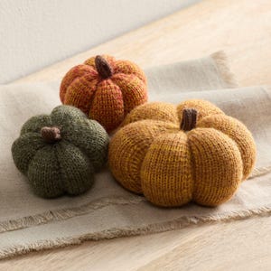 Puede incluir: Tres calabazas tejidas en tonos naranja, verde y amarillo con tallos marrones. Las calabazas están dispuestas sobre una superficie de lino beige, creando un tema otoñal acogedor. La textura tejida añade un toque artesanal a los objetos decorativos.
