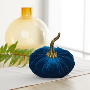 May include: A blue velvet pumpkin with a gold stem. The pumpkin is sitting on a white table runner.