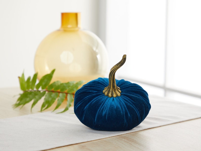 May include: A blue velvet pumpkin with a gold stem. The pumpkin is sitting on a white table runner.