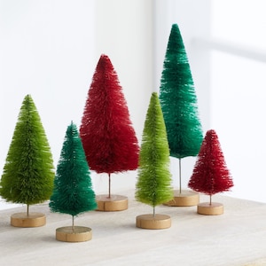 May include: A set of six miniature bottle brush Christmas trees in green and red. The trees are arranged in a row on a wooden surface.