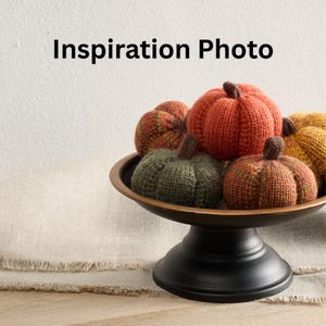 May include: A decorative arrangement of knitted pumpkins in various colors, including orange, green, and yellow, displayed in a bronze-colored bowl on a black pedestal. The text "Inspiration Photo" is at the top.