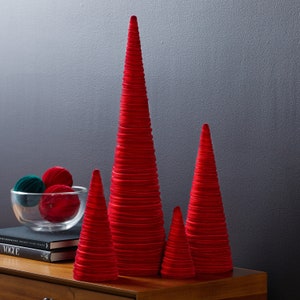 以下が含まれることがあります： 木製の表面に、さまざまなサイズの赤いベルベット製の円錐形のクリスマスツリーが4つ置かれています。ツリーは布製で、重ねられたフリルデザインになっています。