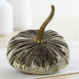 May include: A green velvet pumpkin with a gold stem. The pumpkin is sitting on a white surface.