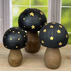 May include: Three black wooden mushrooms with gold star and dot designs. The mushrooms are painted black with gold accents and have a brown wooden base.