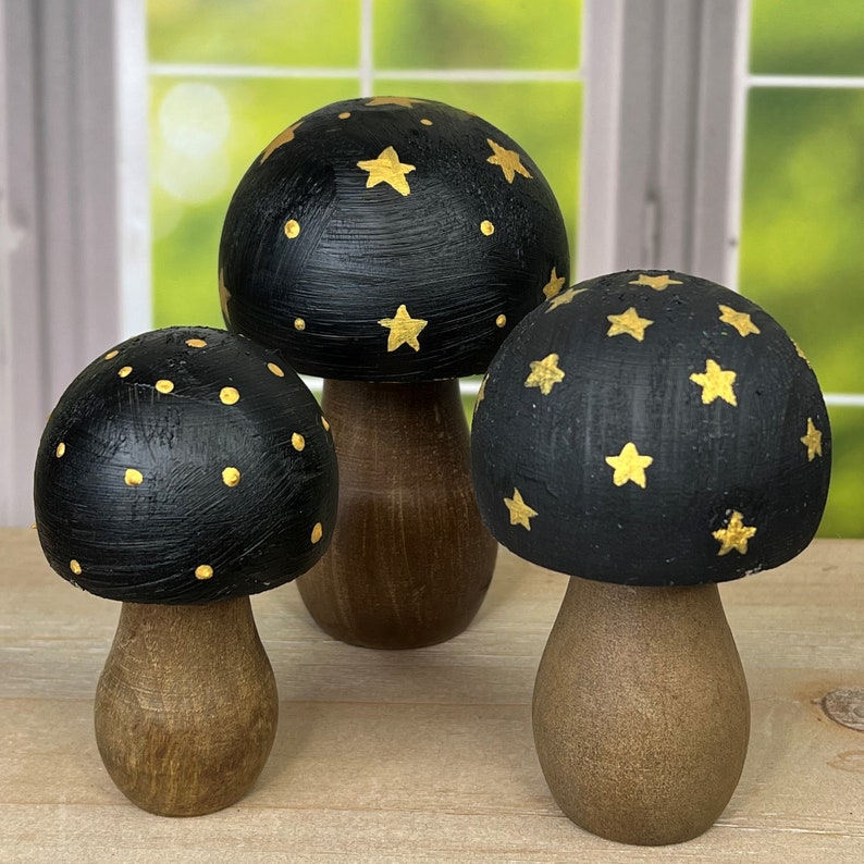 May include: Three black wooden mushrooms with gold star and dot designs. The mushrooms are painted black with gold accents and have a brown wooden base.