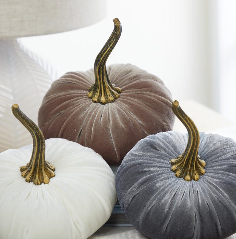 May include: Three velvet pumpkins in white, taupe, and grey. Each pumpkin has a gold-coloured stem. The pumpkins are arranged together, suggesting a fall or harvest theme. The soft texture and colours create a cosy, seasonal aesthetic.