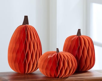 May include: Three orange paper pumpkins with brown stems, in different sizes. The pumpkins have a honeycomb design, creating a textured appearance. They are arranged on a wooden board, ideal for autumn decorations.
