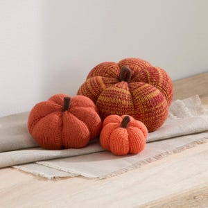 May include: Three knitted pumpkins in varying sizes and shades of orange. The largest pumpkin features a striped pattern of orange and yellow yarn, while the medium and small pumpkins are solid orange. Each pumpkin has a dark brown knitted stem.
