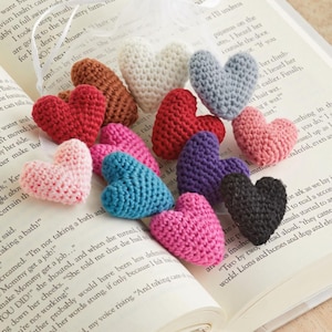 May include: A collection of small, three-dimensional crochet hearts in various colors including white, grey, brown, red, pink, purple, blue, and black. Each heart features a textured, yarn-stitched surface and is gathered together with a white ribbon.