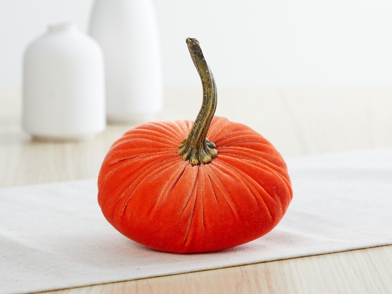 K&ouml;nnte beinhalten: Ein leuchtend orangefarbener Samt-K&uuml;rbis mit einem braunen Stiel. Der K&uuml;rbis liegt auf einer wei&szlig;en Oberfl&auml;che.