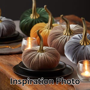 May include: Velvet pumpkins in shades of green, gold, grey, and brown, each with a gold stem, are displayed on a wooden table. Black plates and lit candles add to the autumnal theme. The image includes the text "Inspiration Photo" at the bottom.