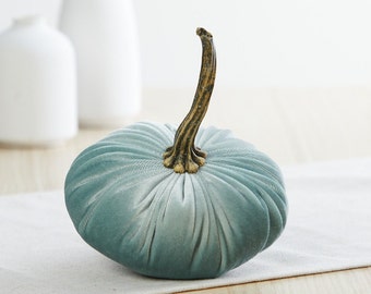 May include: A teal blue velvet pumpkin with a gold stem. The pumpkin is sitting on a white linen surface.