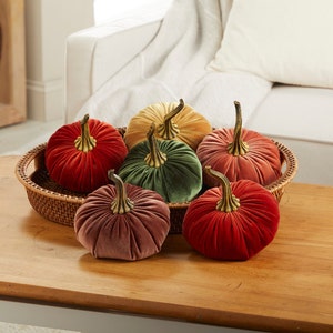 May include: Six velvet pumpkins in various colors, including red, green, pink, and yellow, are arranged in a woven basket on a wooden table. The pumpkins have gold stems and are a decorative fall accent.