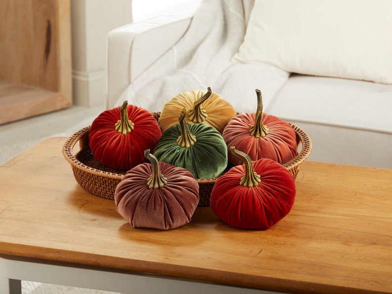 May include: Six velvet pumpkins in various colors, including red, green, pink, and yellow, are arranged in a woven basket on a wooden table. The pumpkins have gold stems and are a decorative fall accent.