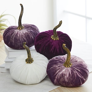 May include: Four velvet pumpkins in shades of purple and white. The pumpkins are arranged on a stack of books and a white tablecloth.