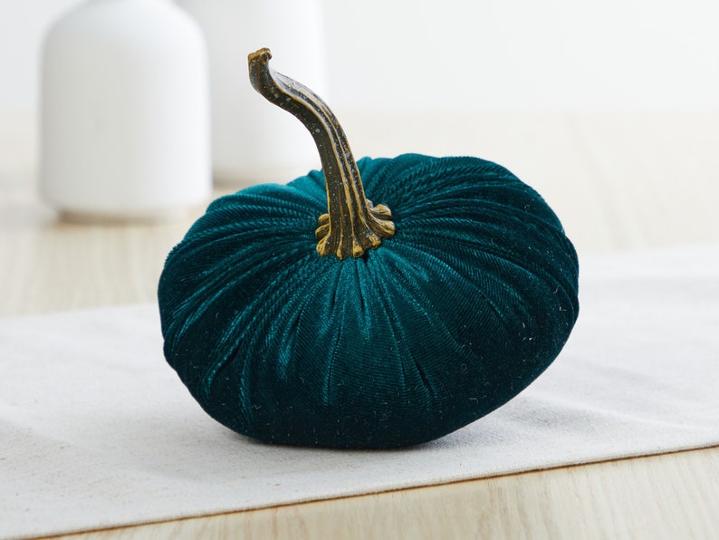 Op de afbeelding: Een teal fluwelen pompoen met een gouden steel. De pompoen ligt op een wit oppervlak.