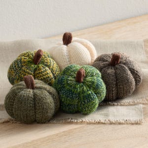 May include: A collection of five knitted pumpkins in various colors, including green, cream, and brown. Each pumpkin has a brown stem and sits on a beige fabric. The pumpkins are arranged on a wooden surface.