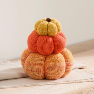 May include: A stack of three knitted pumpkins in varying sizes. The top pumpkin is yellow, the middle is orange, and the bottom is a variegated blend of orange and yellow. Each pumpkin features a ribbed texture and a small dark brown stem on top.