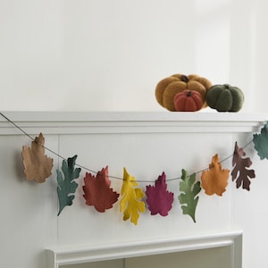 May include: A fall-themed garland of felt leaves in various colors, including brown, green, red, yellow, and purple, hangs in front of a white background. Three decorative pumpkins in orange, green, and gold sit on a shelf above the garland.