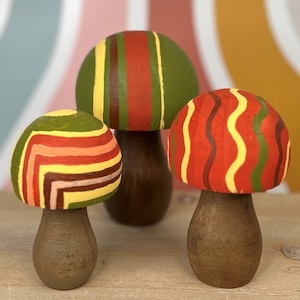 May include: Three wooden mushroom figurines with colorful striped patterns. The mushrooms are painted in shades of red, yellow, and green.