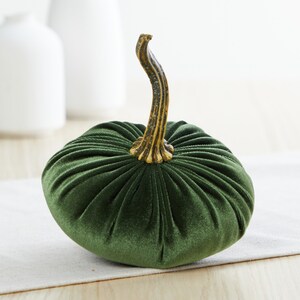 Op de afbeelding: Een groene fluwelen pompoen met een gouden steel. De pompoen staat op een wit tafelkleed.