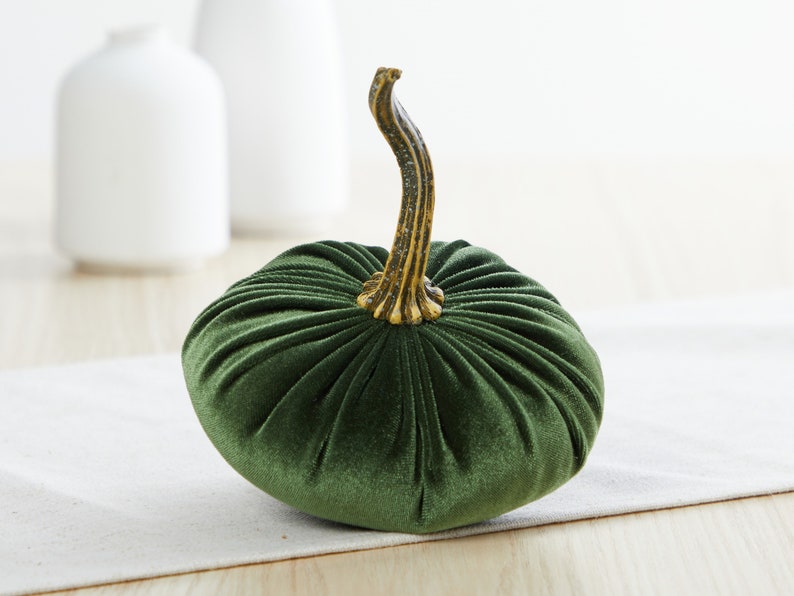 Op de afbeelding: Een groene fluwelen pompoen met een gouden steel. De pompoen staat op een wit tafelkleed.
