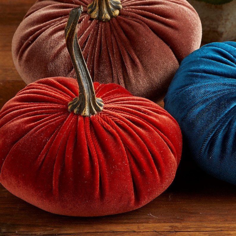 May include: Three velvet pumpkins in shades of red, brown, and blue. The pumpkins are all plump and have a slightly textured surface.