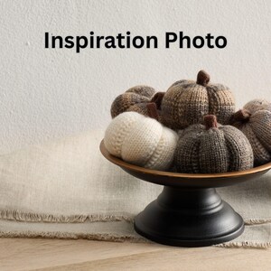 May include: A decorative arrangement of knitted pumpkins in various shades of brown and cream, displayed in a bronze-colored bowl on a black pedestal. The text "Inspiration Photo" is at the top. The pumpkins have brown stems.