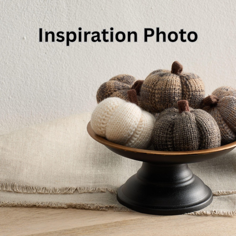 May include: A decorative arrangement of knitted pumpkins in various shades of brown and cream, displayed in a bronze-colored bowl on a black pedestal. The text "Inspiration Photo" is at the top. The pumpkins have brown stems.
