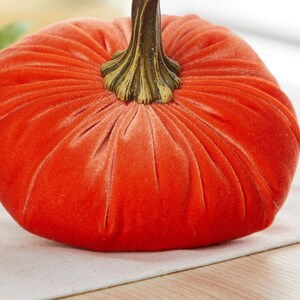 May include: A bright orange velvet pumpkin with a gold-colored stem. The pumpkin is sitting on a white surface.