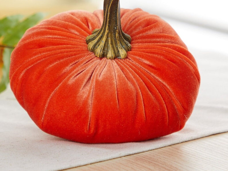May include: A bright orange velvet pumpkin with a gold-colored stem. The pumpkin is sitting on a white surface.