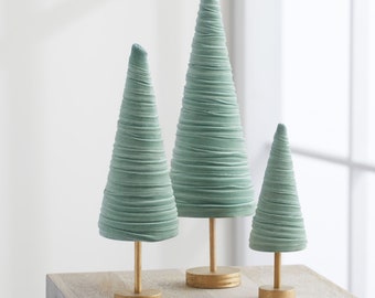 May include: Set of three teal blue cone-shaped Christmas trees with gold bases. The trees are made of a soft, textured material and are arranged in order of size, with the largest tree in the back and the smallest tree in the front.