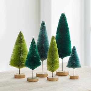 May include: A set of six green and blue bottle brush Christmas trees of varying sizes. The trees are arranged on a light wood surface.