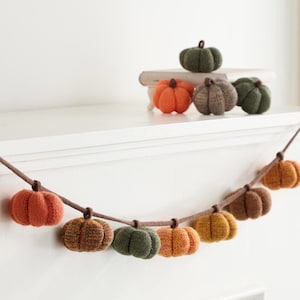 Op de afbeelding: Een decoratieve slinger met een reeks kleine, gebreide pompoenen in tinten oranje, bruin en groen, geregen aan een bruin koord. Extra individuele gebreide pompoenen in vergelijkbare kleuren zijn gerangschikt op een kleine witte plank.