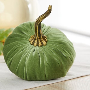 May include: A green velvet pumpkin with a gold and brown stem. The pumpkin is sitting on a white table cloth.