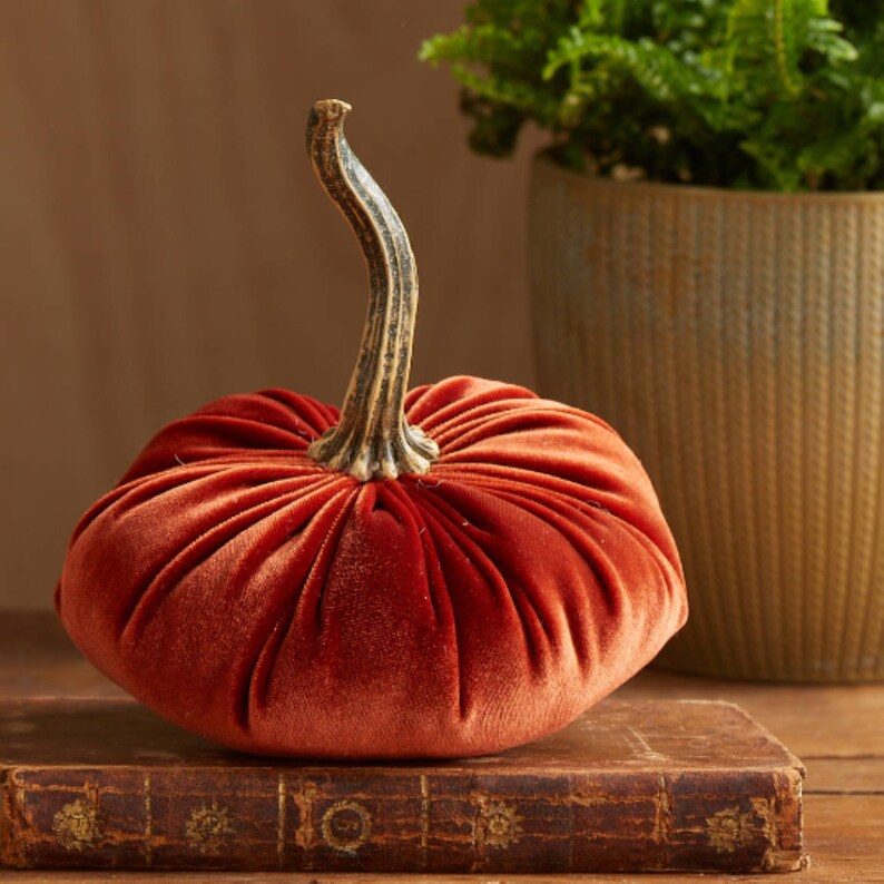 May include: A velvet pumpkin with a gold stem. The pumpkin is a deep orange color and has a pleated design. It is sitting on top of an old book.