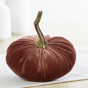 May include: A brown velvet pumpkin with a textured surface. The pumpkin has a curved, brown stem with gold accents. This autumn-themed decoration is suitable for a table or shelf.