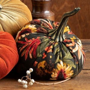Peut inclure: Une citrouille en tissu noir &agrave; motif floral avec une tige brune. La citrouille est pos&eacute;e sur une surface en bois avec d'autres citrouilles en arri&egrave;re-plan.
