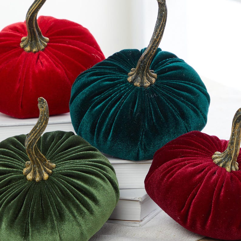 Op de afbeelding: Vier fluwelen pompoenen in rood en groen. De pompoenen hebben een gouden steel en zijn op een wit oppervlak gerangschikt.