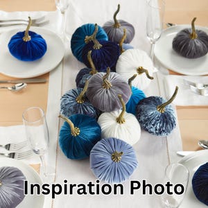 May include: A table setting with a row of velvet pumpkins in shades of blue, gray, and white. The pumpkins are arranged in a line down the center of the table, with a white tablecloth and gold silverware.