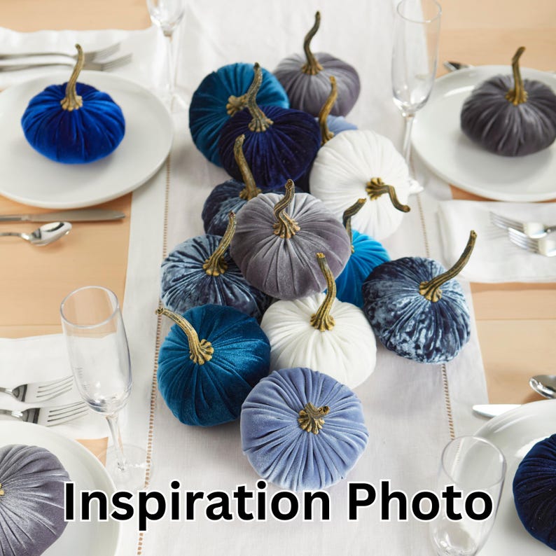 May include: A table setting with a row of velvet pumpkins in shades of blue, gray, and white. The pumpkins are arranged in a line down the center of the table, with a white tablecloth and gold silverware.