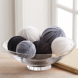 May include: A clear glass decorative bowl filled with decorative fabric spheres in shades of white, gray, and black. The spheres have textured surfaces and are arranged in a decorative display. The bowl sits on a wooden surface.