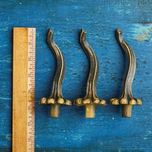 May include: Three decorative metal finials in the shape of a stylised gourd with a brown and gold finish. The finials are arranged horizontally on a blue wooden surface.