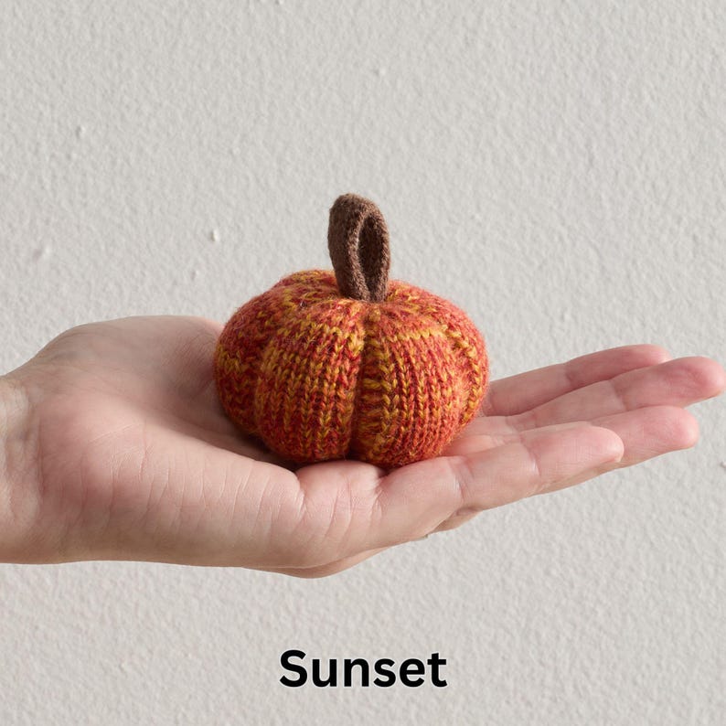 May include: A small, knitted pumpkin in shades of orange and yellow, with a brown stem. The pumpkin is held in a hand, showcasing its size and texture. The word "Sunset" is written in black at the bottom of the image.