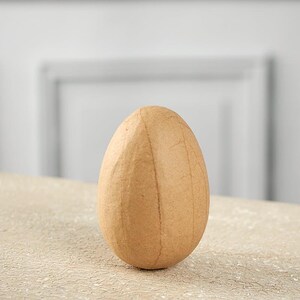 May include: A light brown, egg-shaped object made of paper mache. The object is sitting on a light brown surface.