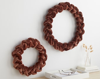 May include: Two brown velvet wreaths, one larger than the other, are hanging on a white wall. The wreaths are made of braided fabric and have a circular shape.