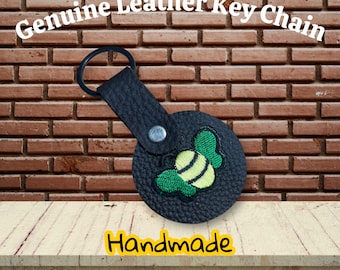 This is a textured black leather snap tab keychain featuring an embroidered green and yellow bee design.