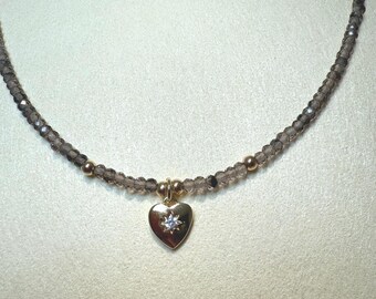 Op de afbeelding: Een delicate ketting met een streng kleine, gefacetteerde rookkwarts kralen. De ketting heeft gouden accenten en een gouden hartvormige hanger met een kleine, sprankelende edelsteen in het midden. De hanger is ongeveer 2 cm breed.