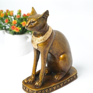 May include: A bronze cat statue in a seated pose, wearing a gold-coloured collar. The statue sits on a rectangular base with a decorative border. The cat has pointed ears and is facing left.