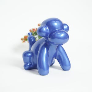 May include: A glossy, blue sculpture of a dog in a seated position. The dog has a rounded body and head, with large ears and a small tail. Small orange flowers are placed in the back. The sculpture is on a white background.